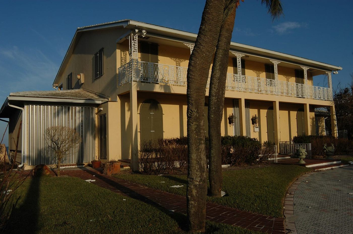 A two-story Florida coastal home with metal storm panels and closed shutters covering its windows and doors, palm trees in the foreground under a clear blue sky