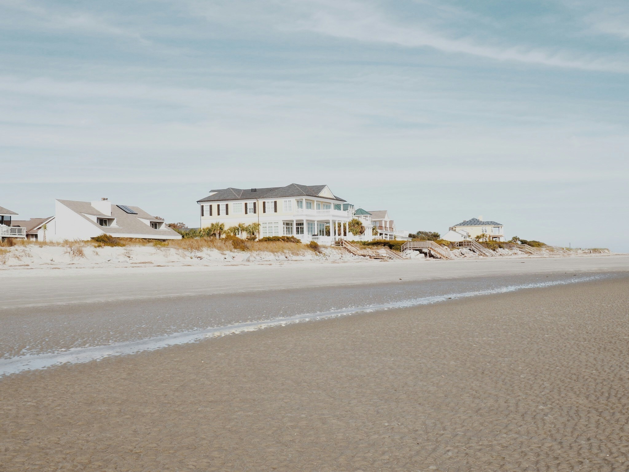An expansive white beachfront home beyond a broad sandy shore. Illustrative architecture, not a current listing.